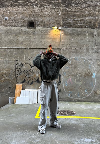 Chaqueta de forro polar verde, pantalones de chándal gris con bolsillos laterales, zapatos tipo slip-on a cuadros y un gorro de rayas multicolores. Fondo urbano.