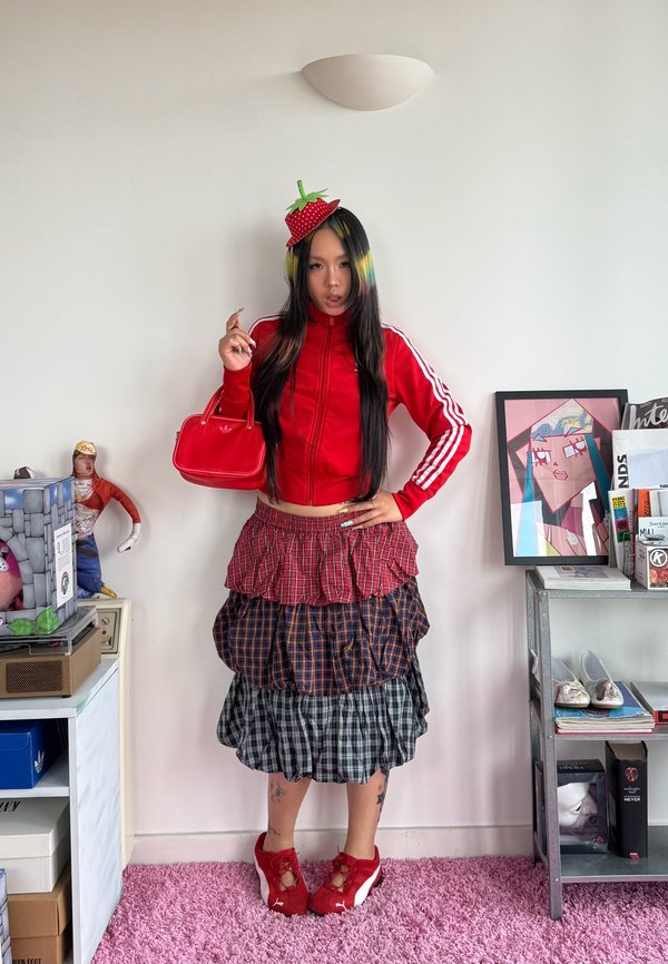 Jonge vrouw in een rode Adidas-jas, gelaagde geruite rok, rode sneakers, houdt een rode handtas vast, draagt een aardbeienhoed, staat in een kamer met kunst en boeken.
