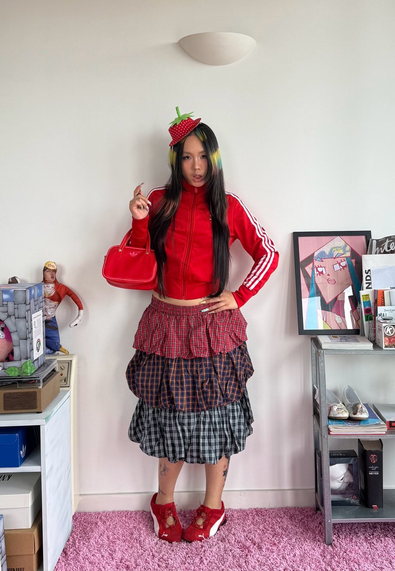 Jeune femme en veste rouge Adidas, jupe à carreaux superposée, baskets rouges, tenant un sac rouge, portant un chapeau fraise, debout dans une pièce avec de l'art et des livres.
