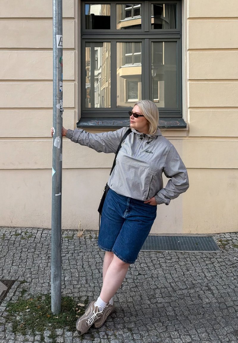 Femme portant des lunettes de soleil, une veste grise et un short en denim, appuyée contre un poteau métallique sur un trottoir pavé près d'une fenêtre d'un bâtiment beige.