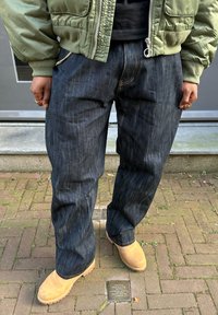 Jeans in denim blu scuro con una vestibilità ampia, cuciture visibili e una cerniera argentata. Abbinati a stivaletti alla caviglia color beige chiaro su una superficie di mattoni.