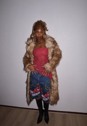 Abrigo de peludo sintético en marrón claro, top de encaje rosa, shorts de denim estampados con diseño floral, y botas negras de tacón alto hasta la rodilla.