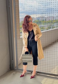 Beige Trenchcoat mit tailliertem Gürtel, schwarzem Spitzenoberteil, schwarzen, knöchellangen Hosen, bordeauxfarbenen spitzen Absätzen und einer gemusterten Handtasche mit Griffen.