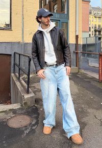 Barna bőrkabát szürke kapucnis pulóver fölött, laza világoskék farmer, és bézs papucsok. A háttérben texturált fal és korlát látható.