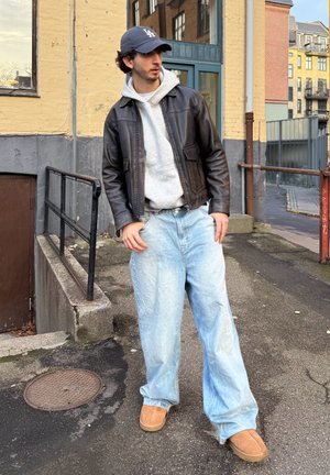 Chaqueta de cuero marrón sobre una sudadera con capucha gris, vaqueros azul claro relajados y zapatillas marrón claro. El fondo presenta una pared texturizada y una barandilla.
