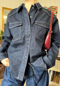 Personne portant une veste en denim bleu foncé et un pantalon assorti, les mains dans les poches, avec un petit sac à bandoulière en cuir rouge.