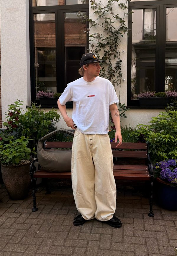 Man wearing white oversized t-shirt, beige wide pants, black cap, standing by wooden bench with plants and windows behind.