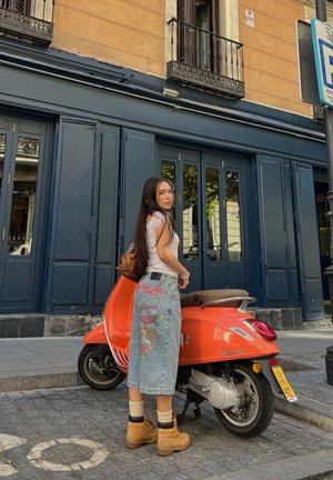 Patinete naranja con un cuerpo redondeado, detalles en plata, estacionado sobre adoquines. Falda de mezclilla con intrincados bordados florales y botas beige.