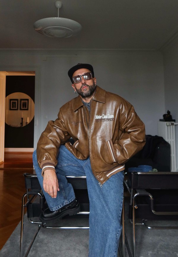 Giubbotto bomber in pelle marrone con cuciture e strisce bianche, abbinato a jeans blu e scarpe nere, ambientato in un interno neutro.