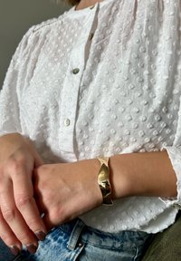 Fehér, texturált blúz kerek gallérral és gombos záródással, sima, arany színű karkötővel egy csupasz csuklón.