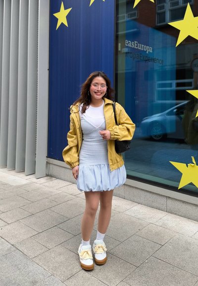 Joven mujer con chaqueta amarilla y vestido azul con rayas está sonriendo junto a una ventana con grandes estrellas amarillas y texto temático de Europa.