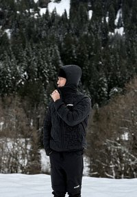 Homme en veste à capuche noire et pantalon, debout dans un paysage enneigé avec une forêt dense d'arbres à feuilles persistantes en arrière-plan, faisant face à gauche.