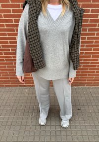 Grå v-ringad tröja med ribbad fåll, lagd över en vit skjorta. Ljusgrå vida byxor och silverfärgade sneakers, med en brun och svart rutig jacka över axlarna.