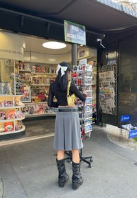 Črna čipkasta bluzica z dolgimi rokavi, siva plisirana krila in črne škornje do kolen z zaponkami. V ozadju je novičarski kiosk z revijami.