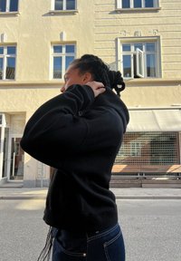 Čierna mikina s kapucňou a textúrovaným povrchom, so prednou kapsou a rebrovanými manžetami, nosená s modrými džínami. Bočný profil.
