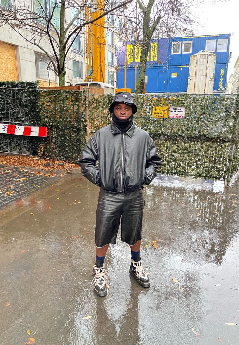 Persona con cappello a secchiello nero, giacca di pelle nera e pantaloni corti di pelle nera che sta in piedi su un pavimento bagnato vicino a un cantiere con una recinzione verde e segnali.