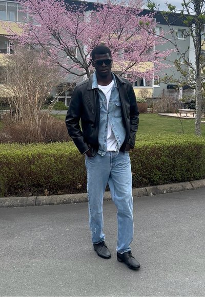 Chaqueta de cuero negra sobre una camisa de denim clara, camiseta blanca y jeans de un azul claro. Zapatos negros. Entorno al aire libre con árboles en flor de color rosa.