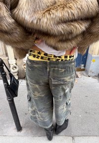 Pantalon cargo camouflage avec détails usés, porté avec une ceinture jaune et noire à logo. Un manteau en fausse fourrure et des bottes noires complètent le look.