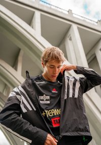 Casaco desportivo preto com riscas brancas nas mangas, usado sobre uma camisa preta com logótipos. Tecido texturizado com um ajuste folgado.