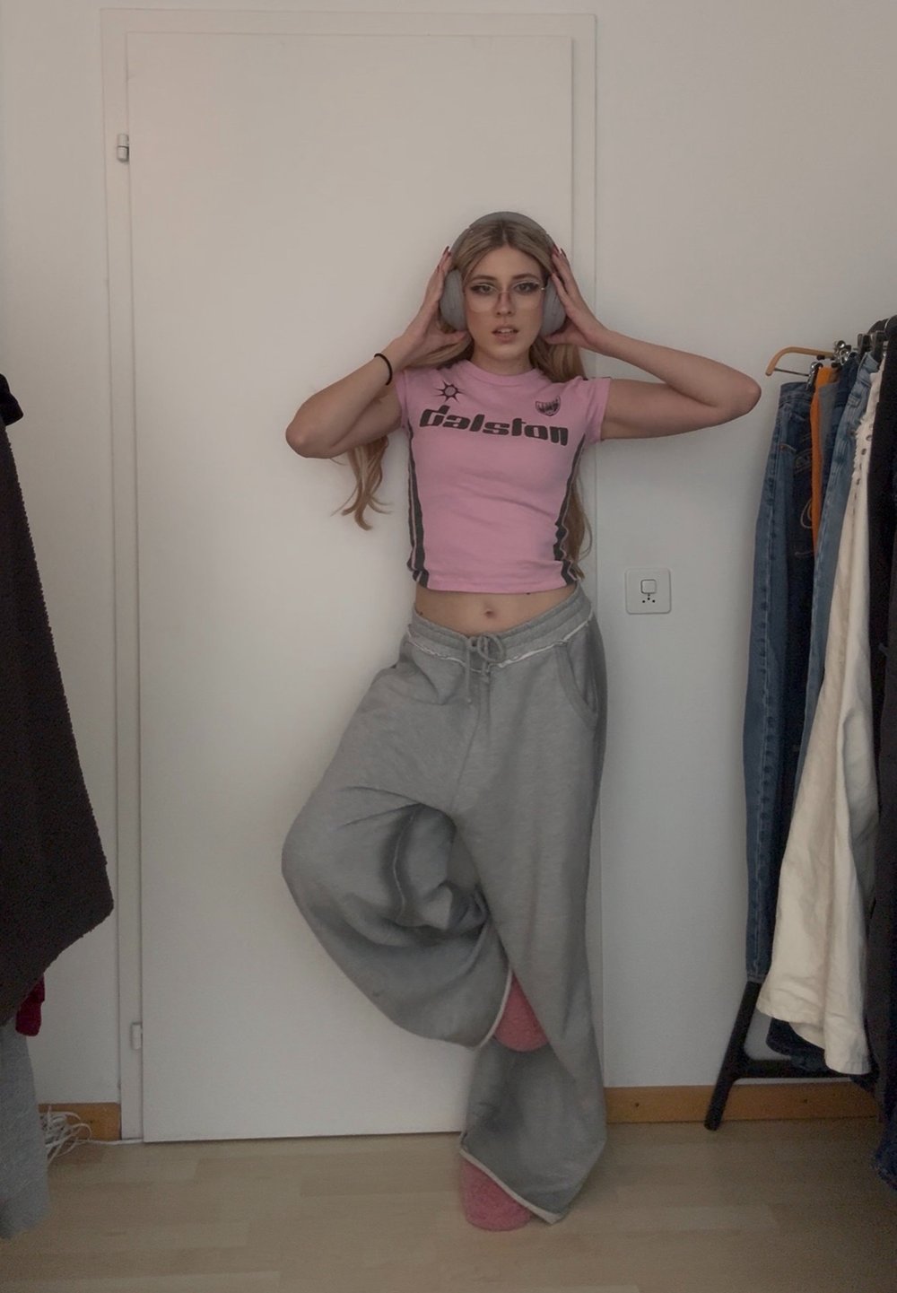 Jeune femme portant des lunettes, un crop top rose, un pantalon de survêtement gris et un casque, debout sur une jambe dans une pièce avec un portant à vêtements.