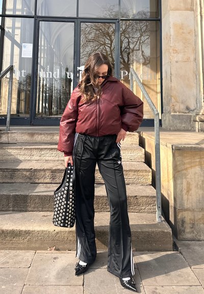 Chaqueta acolchada burdeos con cierre de cremallera, pantalones deportivos negros con rayas laterales, bolso tote negro con tachuelas y zapatos negros de punta.