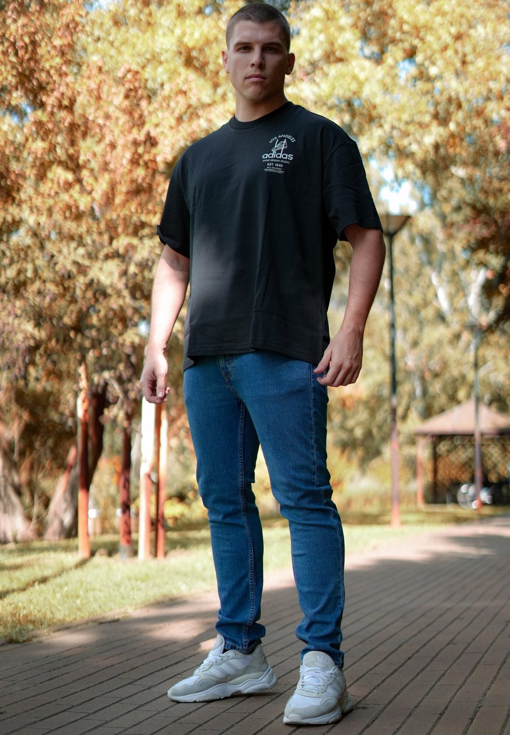 Schwarzes Baumwoll-T-Shirt mit hochgerollten Ärmeln, blaue Jeans und weiße Sportschuhe. Steht im Freien auf einem gepflasterten Weg.