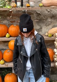 Person i svart läderjacka, grå huvtröja och svart mössa som står framför en utställning av pumpor och gourds på trähyllor.