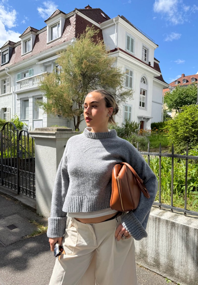 Femme portant un pull gris et un pantalon crème, tenant un sac marron, debout sur un trottoir avec une maison blanche et de la verdure en arrière-plan.