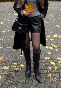 Žena v černém kabátu, kožených šortkách, černých botách a s podzimními listy a černou kabelkou v ruce, stojící na dlážděné cestě s padlými listy.