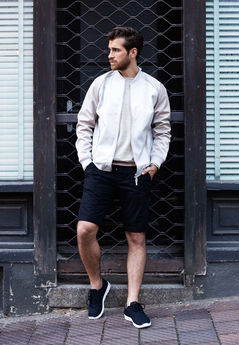 Lichtgrijze bomberjack over een beige trui, zwarte shorts en zwarte sneakers. Man staat tegen een houten achtergrond met een rasterpatroon.