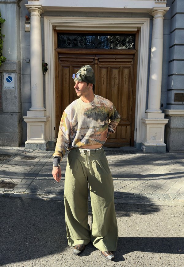 Una persona è in piedi di fronte a porte di legno indossando un maglione multicolore e texturizzato, pantaloni larghi color verde oliva e un berretto verde.