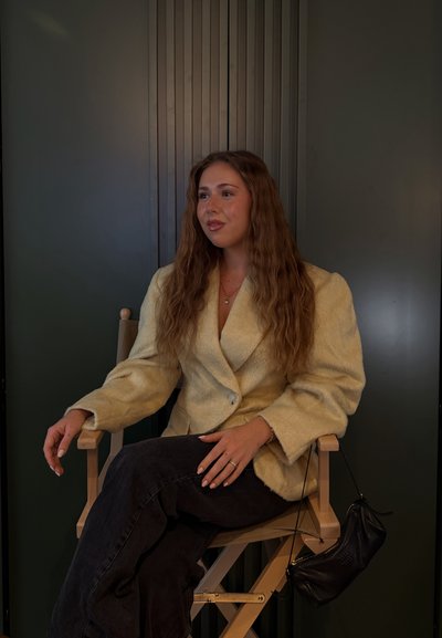 Joven con cabello largo y ondulado sentada con las piernas cruzadas en una silla de madera, vistiendo un blazer beige texturizado y pantalones negros, con un bolso negro.