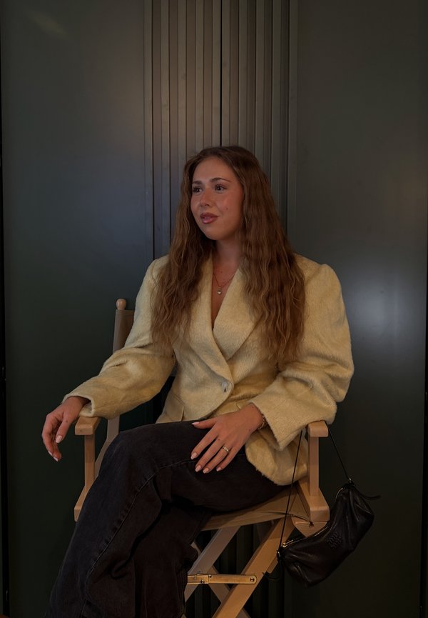 Junge Frau mit langen, gewellten Haaren sitzt im Schneidersitz auf einem Holzstuhl, trägt ein beigefarbenes, strukturiertes Blazer und schwarze Hosen, mit einer schwarzen Handtasche.