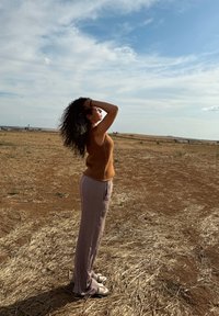 Top senza maniche di maglia marrone, pantaloni a gamba larga a righe beige e marrone, e sandali bianchi. In piedi in un campo asciutto e aperto sotto un cielo blu.