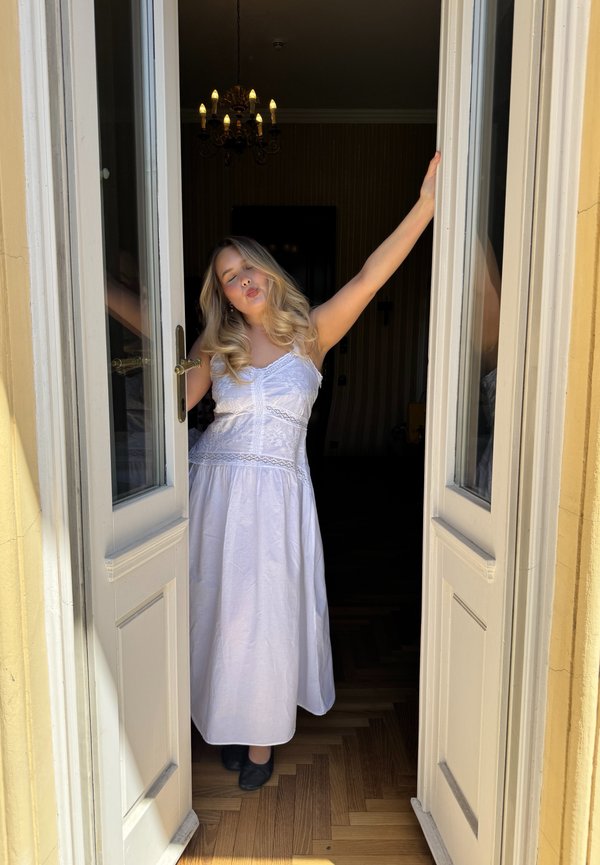 Jeune femme en robe blanche debout dans des portes doubles ouvertes, bras levés, yeux fermés et lèvres pincées à l'intérieur d'une pièce avec un lustre.