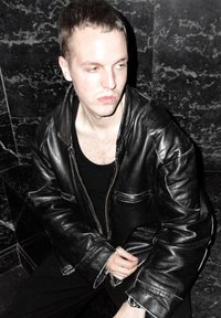 Musta nahkatakki vetoketjulla, jossa on sivutaskut ja näkyvät saumat. Mallilla on päällään musta toppi ja hän istuu tummanmarmorin against.