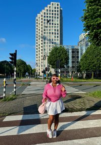 Gaiši rozā adīta džemperis, balta plisētā svārku, rozā saulesbrilles, tur rokā pūkainu rozā somu, stāv uz gājēju pārejas ar mūsdienu ēkām fona.