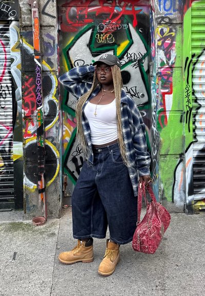 Culottes de mezclilla, camiseta blanca sin mangas, camisa de cuadros oversize, botas de trabajo beige y un bolso rojo texturizado frente a un colorido fondo de graffiti.