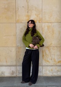 Overdimensioneret grøn strikket turtleneck sweater, sorte brede bukser og brun ruskind handbag, mod en struktureret beige væg.