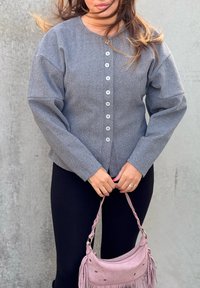 Cardigan gris à motif chevrons avec une patte de boutonnage, manches longues et coupe décontractée, accompagné d'un petit sac à franges rose.