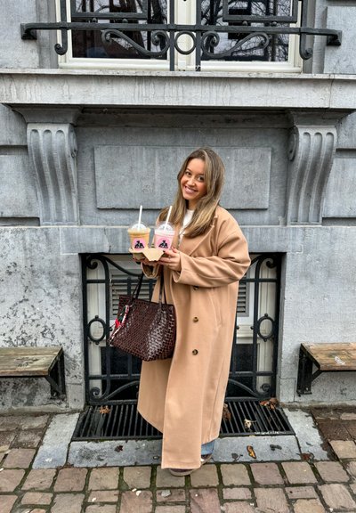 Abrigo beige de gran tamaño, bolso marrón con patrón de cocodrilo, sosteniendo dos bebidas en vasos transparentes con bebidas rosas y beige, acentos negros en los vasos.
