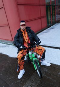 Világos narancssárga melegítő, absztrakt fekete és rózsaszín mintákkal, fekete steppelt dzsekivel párosítva, zöld kerékpáron ülve havas területen.