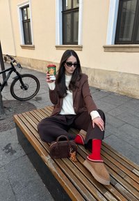 Jeune femme portant des lunettes de soleil, un costume marron, des chaussettes rouges et des chaussures beiges, assise sur un banc, tenant une tasse festive, avec un sac marron et un vélo noir à proximité.