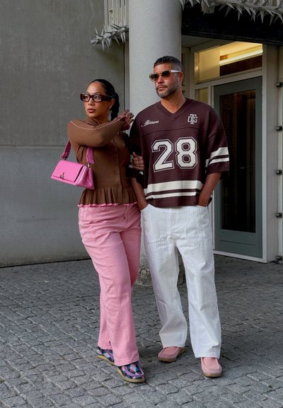 Top peplum marrón, pantalones rosas y bolso rosa en una persona; la otra lleva una camiseta gráfica marrón y pantalones blancos con zapatos estampados.