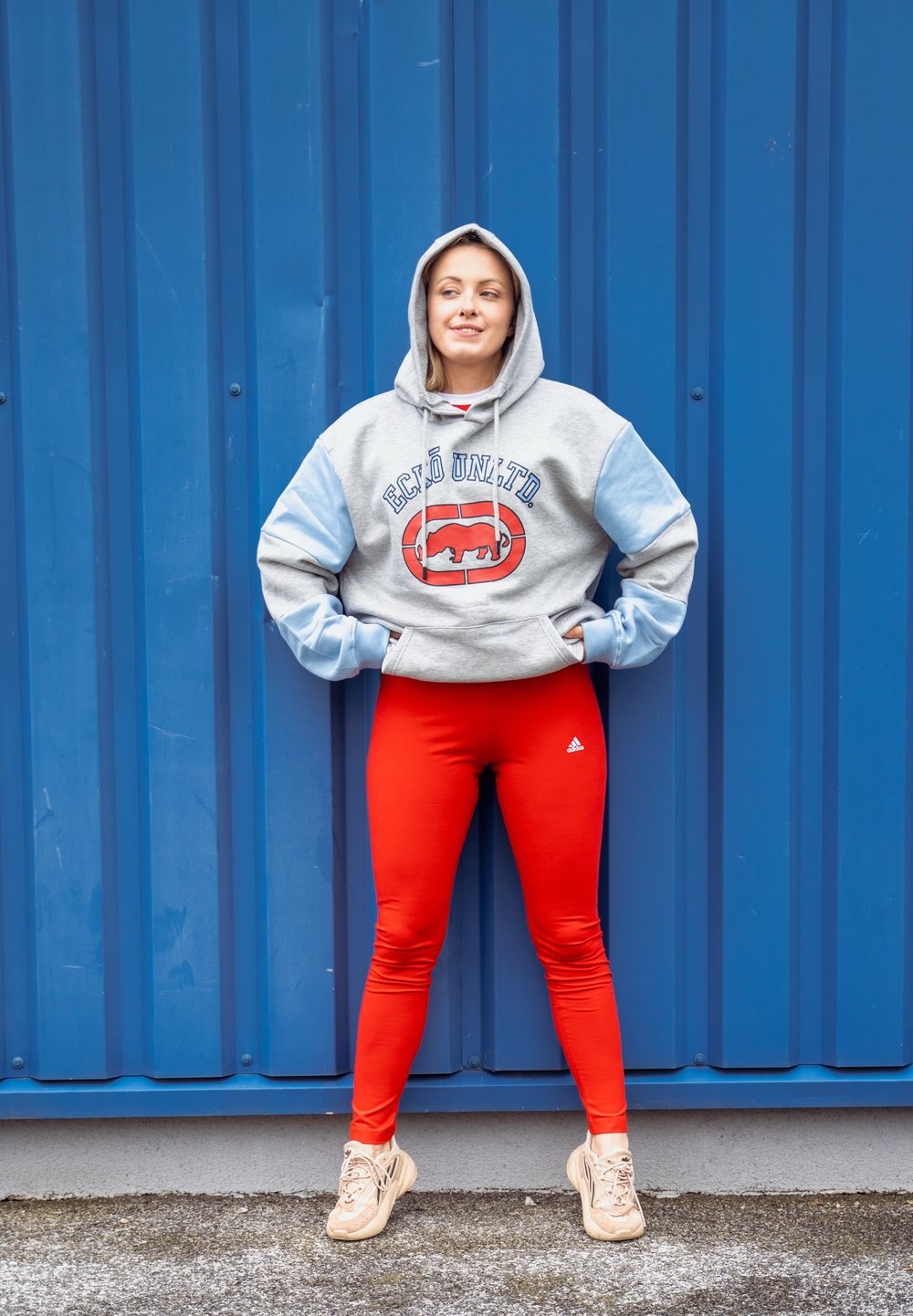 Γκρίζα και ανοιχτό μπλε φούτερ σε μεγάλη διάσταση με λογότυπο, συνδυασμένο με φωτεινές κόκκινες κολάν και μπεζ αθλητικά παπούτσια, standing στην μπλε background.