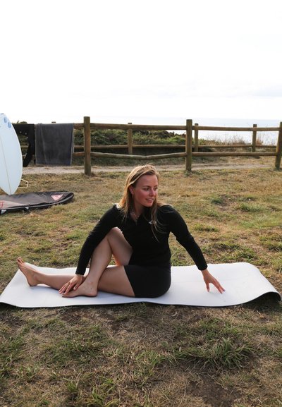 Esterilla de yoga en gris claro sobre el césped, con una persona en una camiseta de manga larga negra y pantalones cortos realizando un estiramiento sentado. En el fondo, tablas de surf.