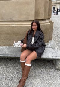 Fiatal nő barna csizmában és bőrkabátban egy kőpadon ül, kezében tálca két elviteles kávéval és harapnivalóval, odakint.