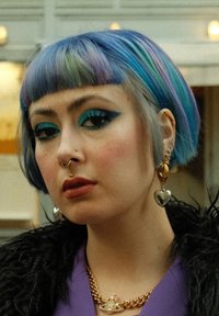 Coupe de cheveux bob courte avec des reflets bleus et violets. Maquillage des yeux bleu audacieux, boucles d'oreilles en or et en argent, et un collier en chaîne dorée épaisse.