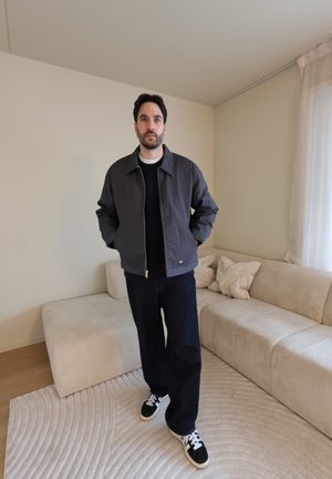 Hombre de pie en una sala de estar beige, vestido con una chaqueta oscura, pantalones negros y zapatillas blancas y negras, con las manos en los bolsillos.