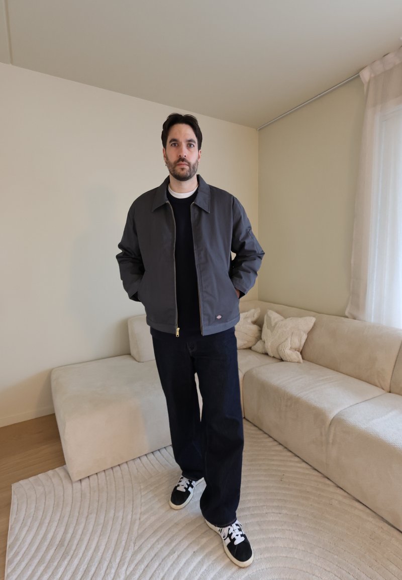 Homme debout dans un salon beige portant une veste sombre, un pantalon noir et des baskets noires et blanches, les mains dans les poches.